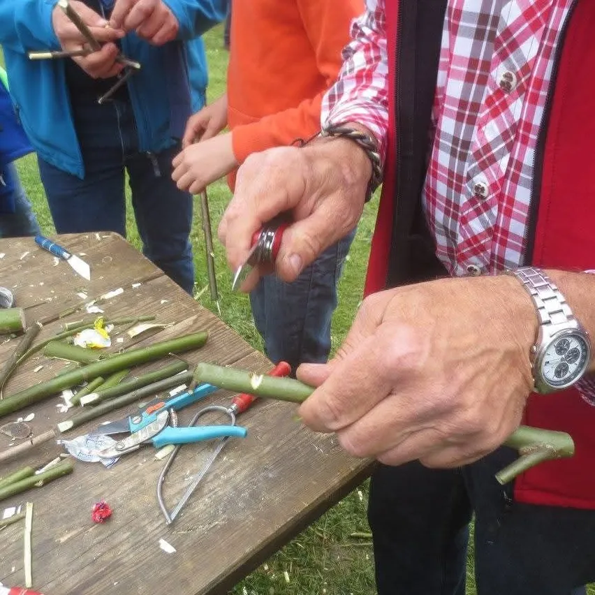 Eine Person demonstriert eine Stockschnitztechnik mit einem Messer. Ein Tisch hat mehrere Stöcke und Gartenwerkzeuge. Andere Personen stehen um den Tisch herum.