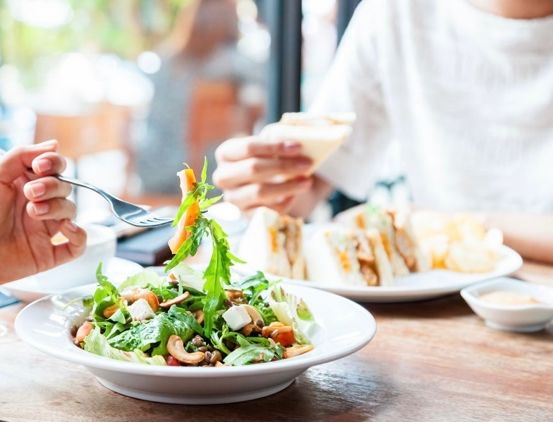 Zwei Personen essen in einem Restaurant. Eine hält einen Salat mit einer Gabel.