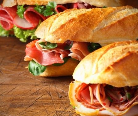 Drei Sandwiches mit verschiedenen Fleischsorten und Gemüsen sind auf einem Holztisch angeordnet, wobei der Fokus auf Frische und appetitlicher Präsentation liegt.