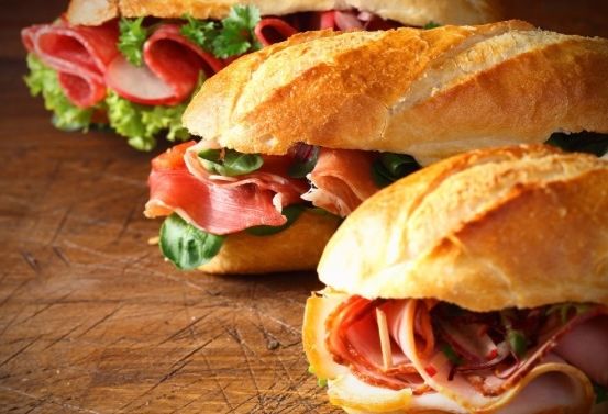 Drei Sandwiches mit verschiedenen Fleischsorten und Gemüsen sind auf einem Holztisch angeordnet, wobei der Fokus auf Frische und appetitlicher Präsentation liegt.