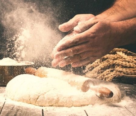 Eine Person knetet Teig auf einem Holztisch, um sie herum verstreut Mehl und ein Nudelholz in der Nähe. Ein Behälter mit Mehl steht links.