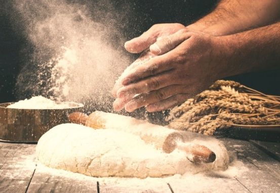 Eine Person knetet Teig auf einem Holztisch, um sie herum verstreut Mehl und ein Nudelholz in der Nähe. Ein Behälter mit Mehl steht links.