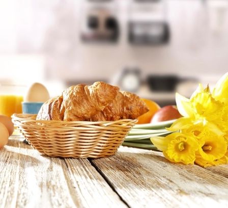 Ein Holztisch mit einem Korb mit Croissants, Eiern und gelben Blumen. Ein Glas Orangensaft und eine blaue Schüssel sind links.