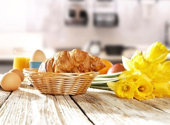 Ein Holztisch mit einem Korb mit Croissants, Eiern und gelben Blumen. Ein Glas Orangensaft und eine blaue Schüssel sind links.