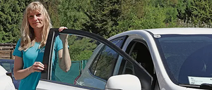 Eine Frau mit blonden Haaren steht neben einem weißen Auto mit schwarzen Schriftzug, 'Caritas & Du' an der Seite. Sie trägt ein blaues T-Shirt und graue Hose. Hinter ihr sind Bäume und ein Gebäude.