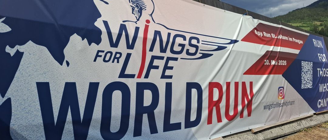 Ein Banner mit einer Silhouette eines laufenden Menschen, der Text 'WINGS FOR LIFE WORLD RUN' in fetten blauen und roten Buchstaben auf weißem Hintergrund.
