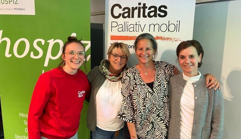 Vier Frauen stehen lächelnd vor einem Banner mit Caritas und mobiler Palliativversorgung darauf geschrieben. Sie lächeln und scheinen für ein Foto zu posieren.