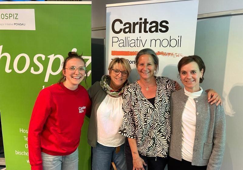 Vier Frauen stehen lächelnd vor einem Banner mit Caritas und mobiler Palliativversorgung darauf geschrieben. Sie lächeln und scheinen für ein Foto zu posieren.