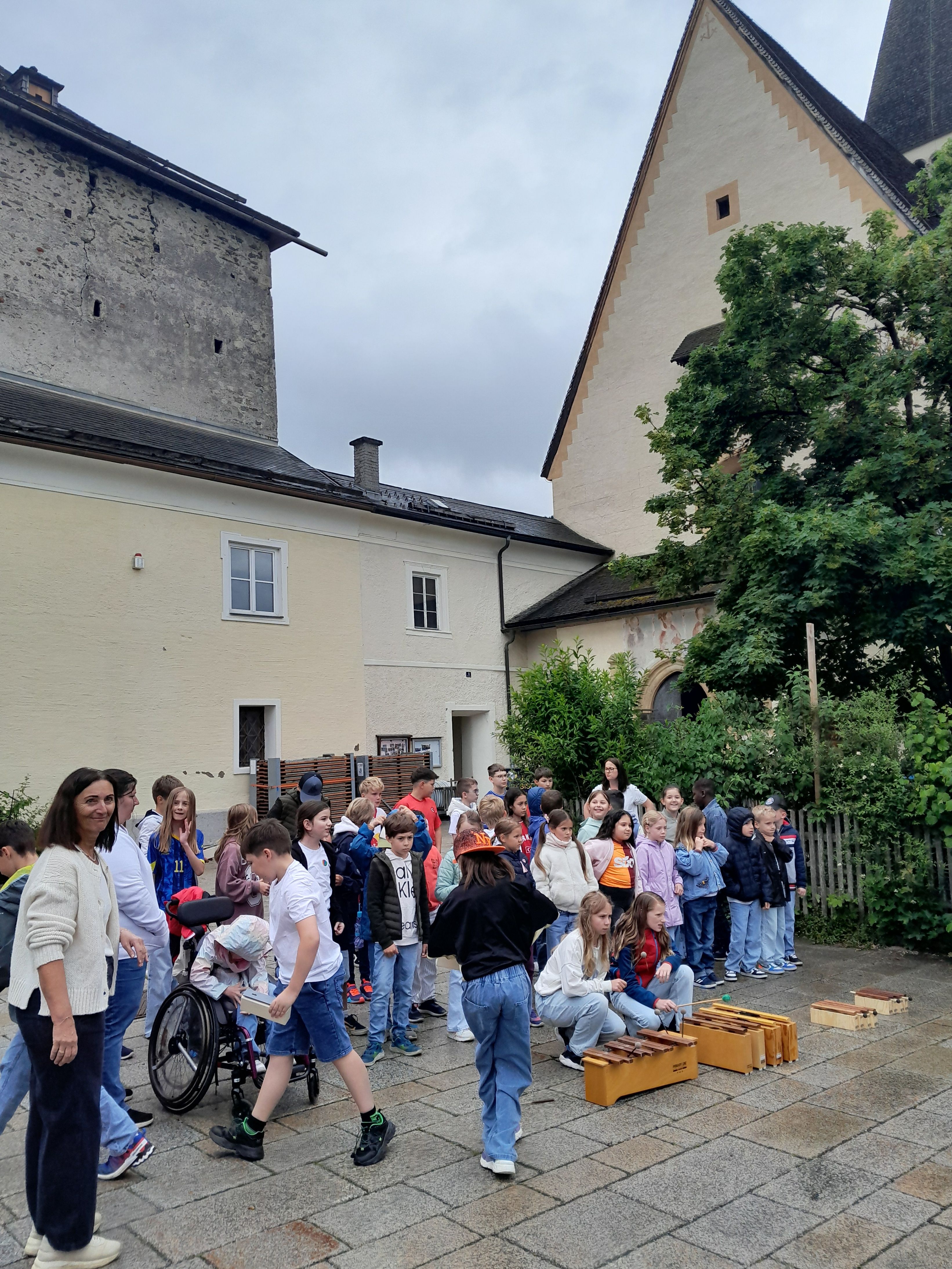 Eine große Gruppe von Kindern und Erwachsenen versammelt sich vor einem Gebäude, einige sitzen auf dem Boden und spielen Xylophon, andere stehen oder sitzen vor einer Frau im Rollstuhl.