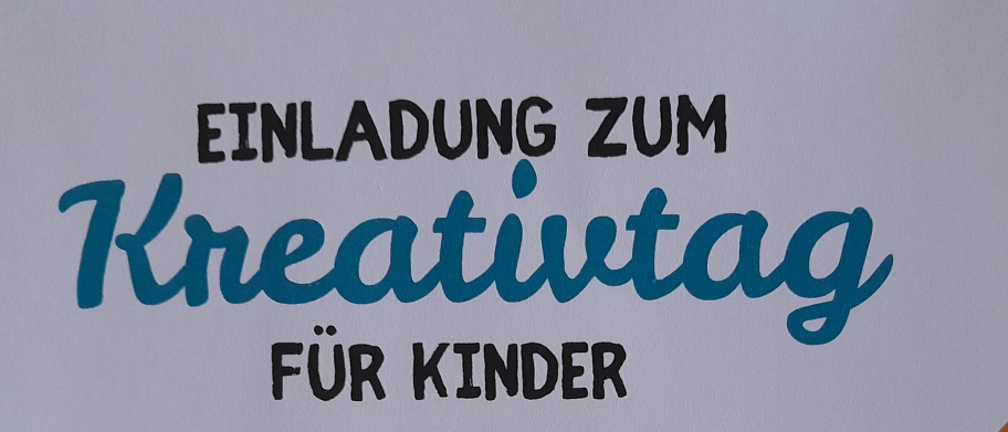 Ein weißes Blatt Papier mit blauem Text lautet 'Einladung zum Kreativtag für Kinder.' Die Worte sind in einer verspielten, kursiven Schriftart geschrieben.