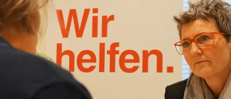 Eine Frau mit Brille sieht eine andere Person ernst an. Hinter ihr steht ein weißes Schild mit roten Buchstaben, das 'Wir helfen' sagt.