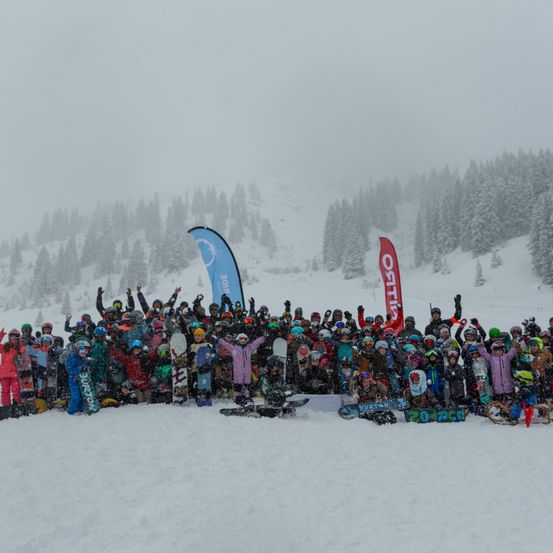 Eine Gruppe von Snowboardern steht in einem verschneiten Bereich und posiert für ein Foto mit Snowboards und Bannern im Hintergrund.