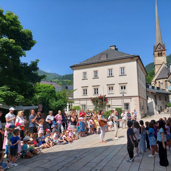 Eine Gruppe von Kindern und Erwachsenen versammelt sich vor einem Gebäude mit einem Turm, und hört einer Frau zu, die eine Gitarre spielt. Das Gebäude ist von Bäumen umgeben und Berge sind im Hintergrund zu sehen.