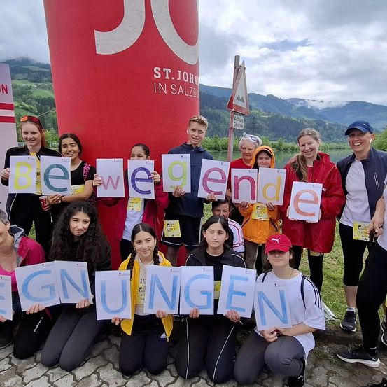 Eine Gruppe von Personen posiert für ein Foto und hält Schilder, die das Wort 'bewegende' bilden. Hinter ihnen steht ein roter Banner mit 'ST. JOHANN IN SALZBURG'. Sie befinden sich auf einem Rasengelände in der Nähe eines Sees mit Bergen im Hintergrund.