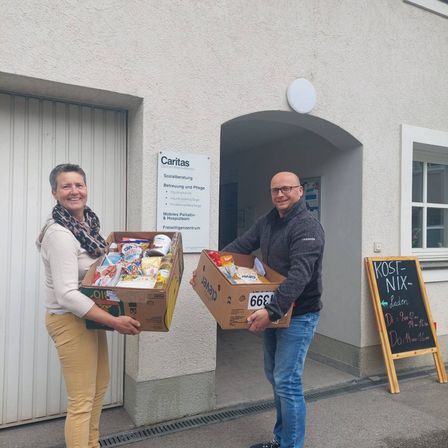 Ein Mann und eine Frau stehen vor einem Gebäude mit einem Schild, das Caritas liest, und halten Lebensmittelboxen.