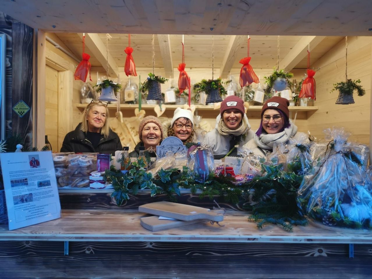 Fünf lächelnde Frauen in einer dekorierten Holzhütte voller Feiertagsartikel und Geschenke. Regale mit hängenden Dekorationen und einem Holztisch mit Lebensmitteln und einem Schild.
