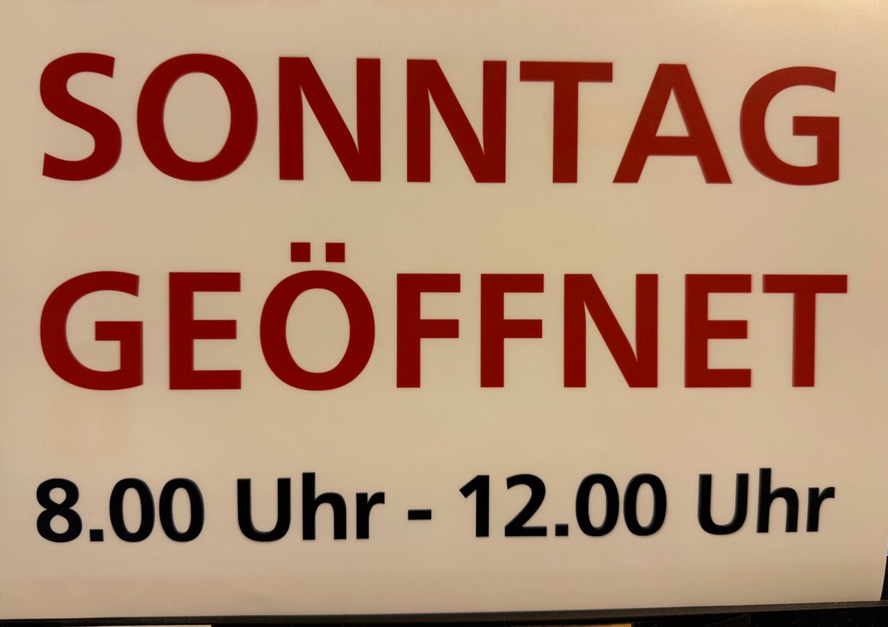 Ein weißes Schild mit rotem Text lautet 'ONNTAGEOEFFNE 00 UHR - 12.00 U'.