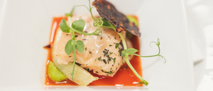 Ein weißer Teller mit einem Fischstück, das mit Kräutern und Gemüse garniert ist und von einer roten Sauce umgeben ist.