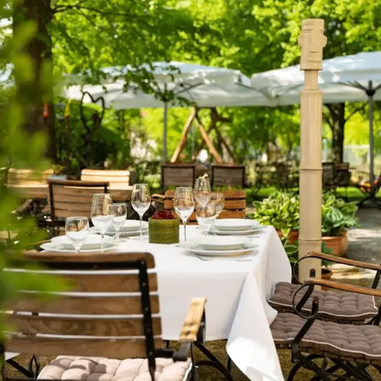 Ein Gartenrestaurant mit eingerichteten Tischen zum Essen. Teller, Weingläser und ein Obstkorb schmücken den Tisch. Sonnenschirme spenden Schatten, und Bäume umgeben den Bereich.