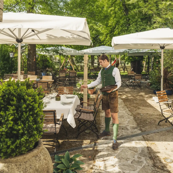 Ein Kellner in grüner Weste und traditioneller deutscher Kleidung serviert einen Tisch in einem Außenrestaurant. Der Tisch ist mit Tellern, Weingläsern und einer Schüssel gedeckt.