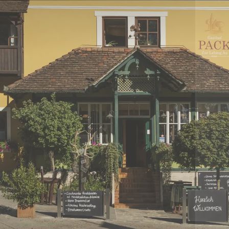 Die Vorderseite eines traditionellen gelben Gebäudes mit einem gefliesten Dach und grünem Zierrat. Draußen befinden sich Schilder in deutscher Sprache und Pflanzen und Bäume schmücken den Eingang. Eine Terrasse ist im oberen Stockwerk sichtbar.