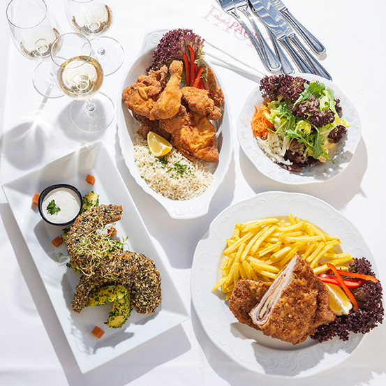 Ein Tisch mit drei weißen Tellern mit Essen, einer mit gebratenem Hähnchen, einem mit Salat und einem mit Pommes. Auch Weingläser und Besteck sind vorhanden.