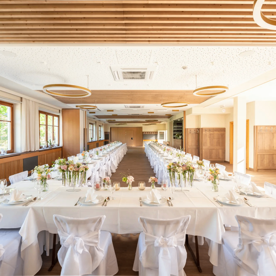 Ein langer Bankettsaal mit weißen Tischen und Stühlen, Holzschränken und großen Fenstern. Die Tische sind mit Blumen, Tellern und Besteck gedeckt.