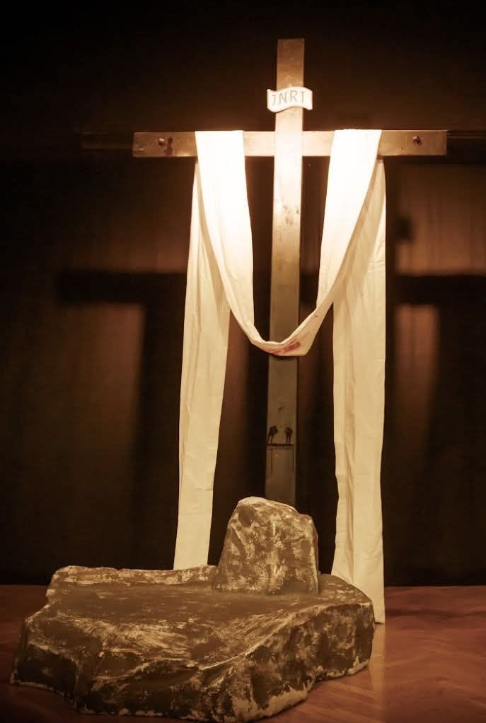 Ein hölzernes Kreuz mit INRI oben steht in einem schwach beleuchteten Raum. Ein weißer Stoff ist über das Kreuz gelegt. Ein Felsblock ist an der Basis platziert.