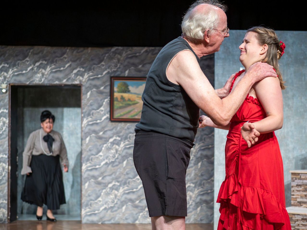 Ein älterer Mann und eine Frau tanzen auf der Bühne, mit einer Frau im Hintergrund.