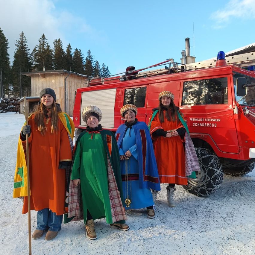 Vier Personen in farbenfrohen Kostümen stehen vor einem roten Feuerwehrfahrzeug. Sie alle lächeln und scheinen für ein Foto zu posieren.