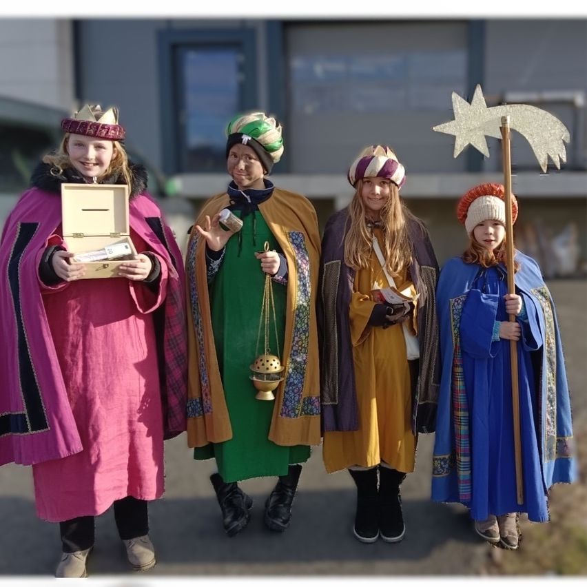 Vier Kinder in farbenfrohen Kostümen, möglicherweise für ein Weihnachtsspiel, stehen zusammen draußen. Eines hält einen Stern, ein anderes eine Laterne, während andere kleine Schachteln und einen Stab tragen.
