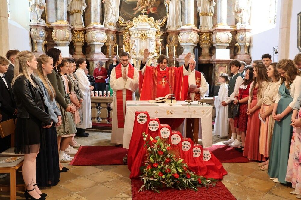 Zwei Priester stehen vor einem Altar, einer trägt eine Brille. Sie sind von Menschen umgeben, einige in formeller Kleidung, in einer Kirche. Der Altar hat Blumen, Kerzen und ein Buch.