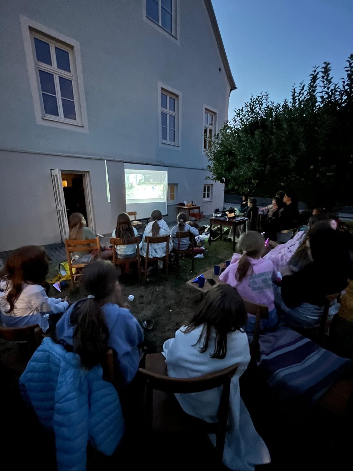 Eine Gruppe von Menschen schaut sich draußen vor einem Gebäude bei Nacht einen Film an. Sie sitzen auf Stühlen, die in einem Kreis angeordnet sind.