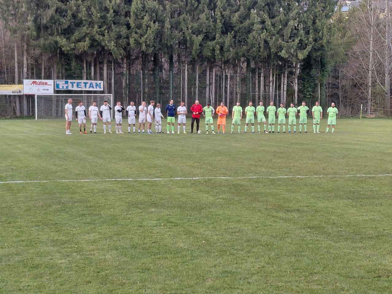 A soccer team lines up on a field, divided into two groups. They wear white and green uniforms. The field is surrounded by tall trees.