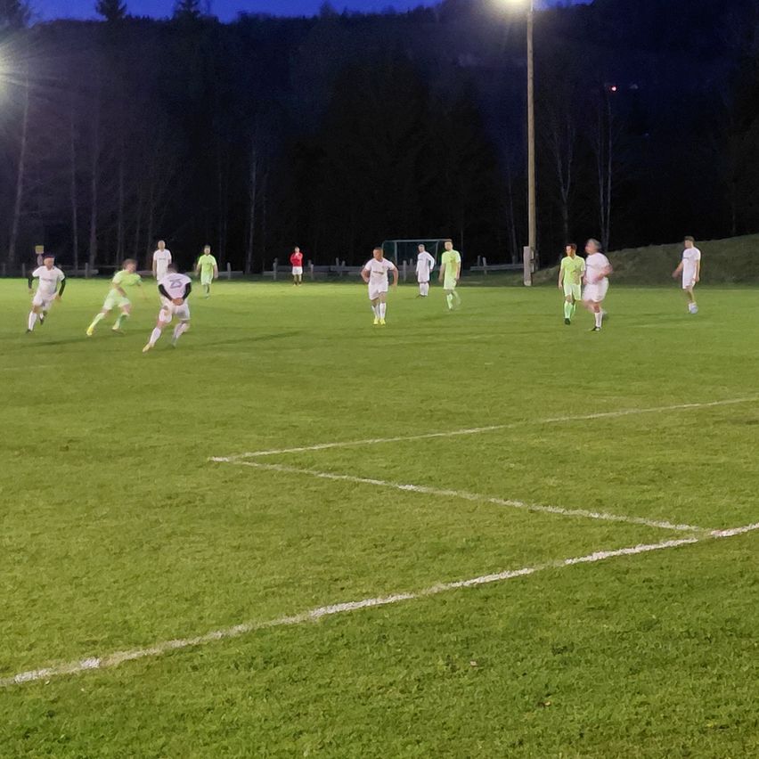 At night, soccer players are playing on a field with white markings. The players are wearing green jerseys. Trees and mountains are in the background.