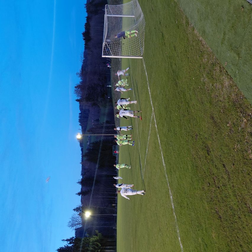 An aerial view of a soccer field shows players in green and white jerseys. One team is near the goal, while the other is positioned further back.