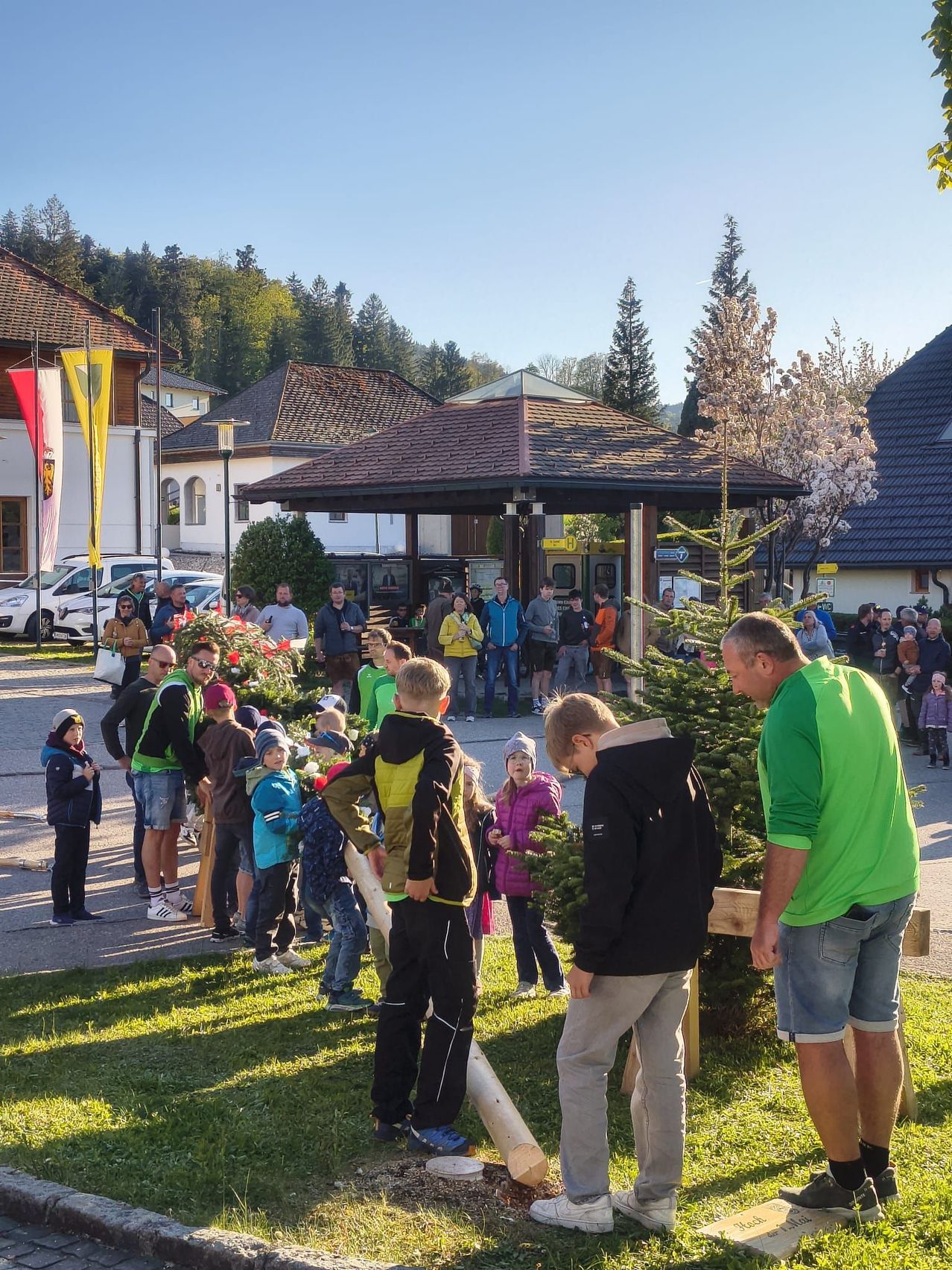 Eine Gruppe von Menschen versammelt sich im Freien, einige stehend und andere gehend, vor einer kleinen Struktur mit einem braunen Dach. Ein Mann hält einen Topf mit einem Baum. Eine Menschenmenge schaut zu.