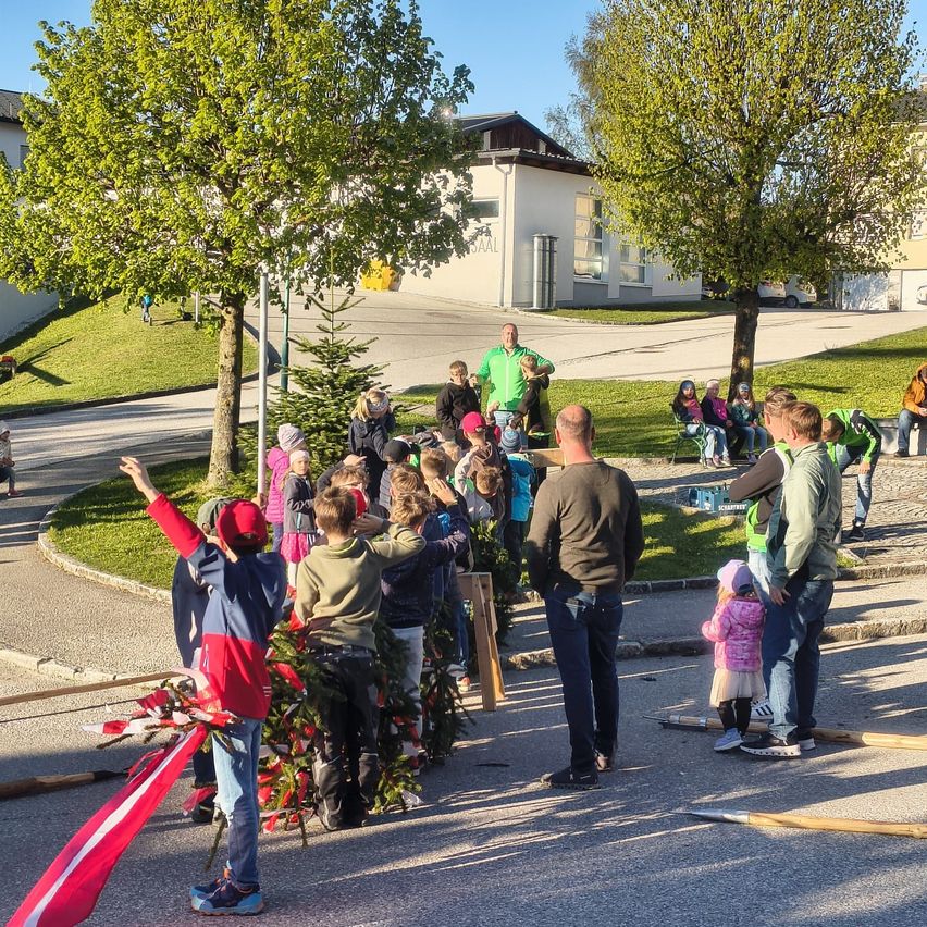 Eine Gruppe von Kindern und Erwachsenen steht um einen Weihnachtsbaum herum, möglicherweise in einem Park. Sie sind in festlicher Kleidung gekleidet. Ein Mann wedelt mit einer Flagge, während eine Frau ein Kind trägt. Im Hintergrund befinden sich Häuser und ein Auto.