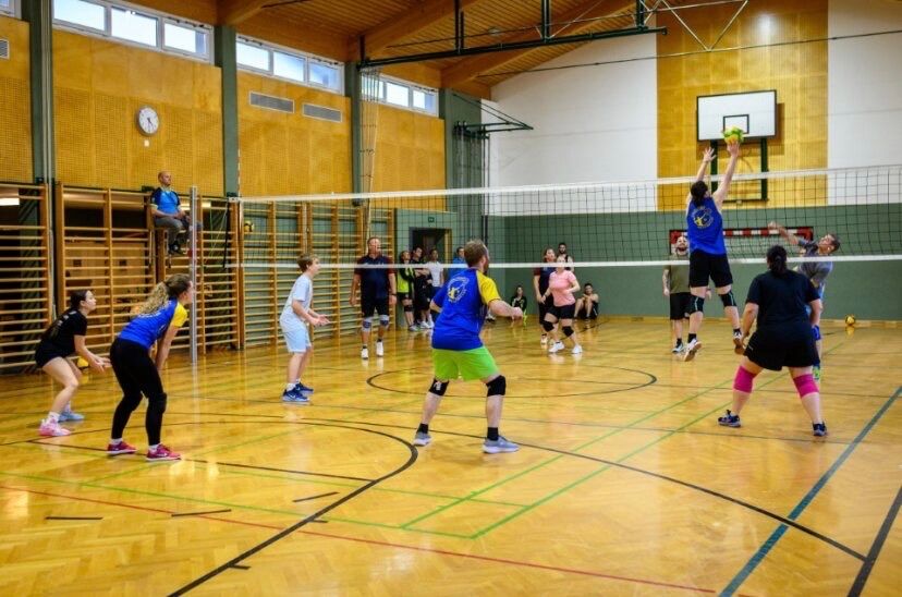 Eine Gruppe von Menschen spielt Volleyball in einer Turnhalle. Das Netz teilt den Platz, und die Spieler sind in Aktion. Ein Spieler springt, um den Ball zu schlagen. Andere schauen zu und warten auf ihren Zug.