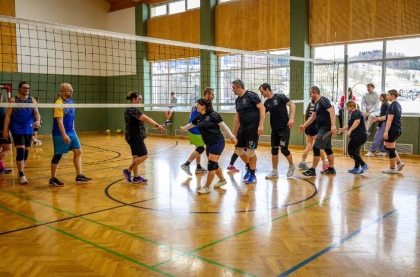 Eine Gruppe von Menschen, sowohl Männer als auch Frauen, spielen Volleyball in einer Halle. Sie tragen alle schwarze T-Shirts und Shorts.
