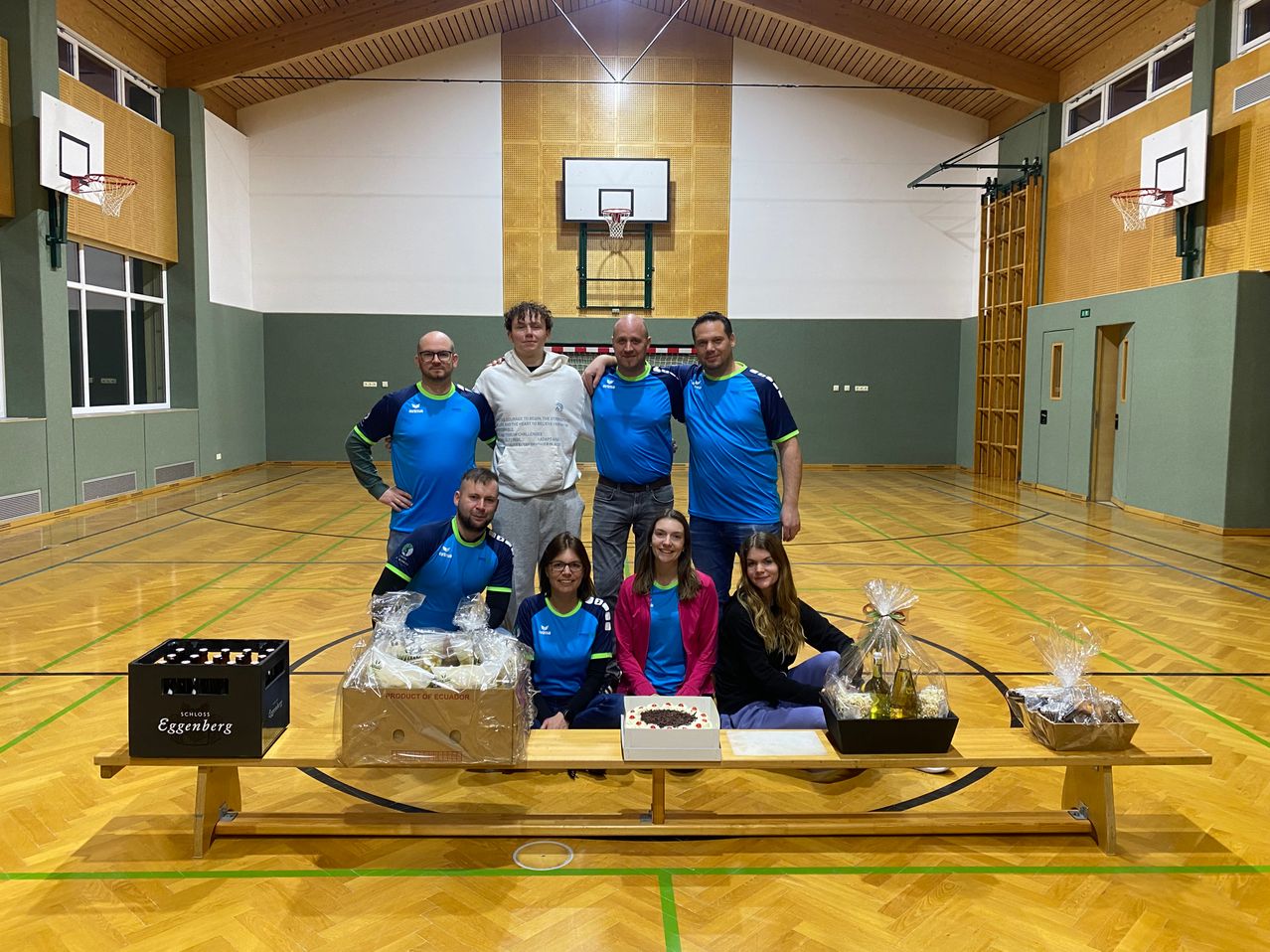 Eine Gruppe von Menschen in Sportkleidung posiert für ein Foto in einer Turnhalle. Sie sind von Geschenkkörben, einem Kuchen und einer Produktbox umgeben. Im Hintergrund ist der Basketballkorb zu sehen.