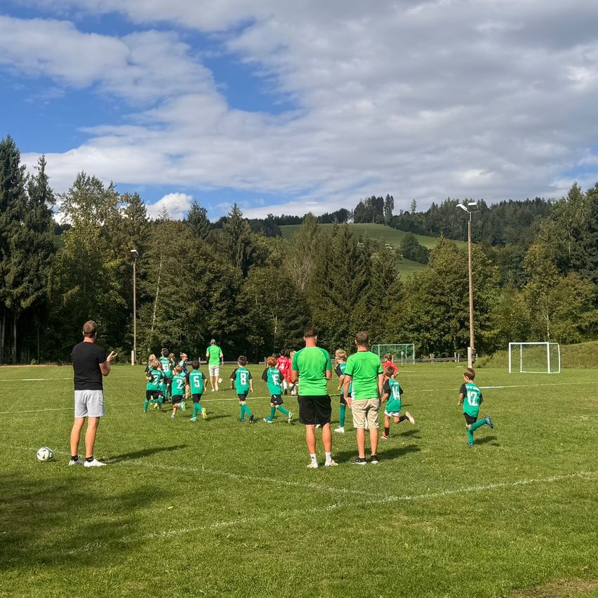 Eine Gruppe junger Fußballspieler in grünen Trikots trainiert auf einem Feld mit einem Trainer und einem Fußball.