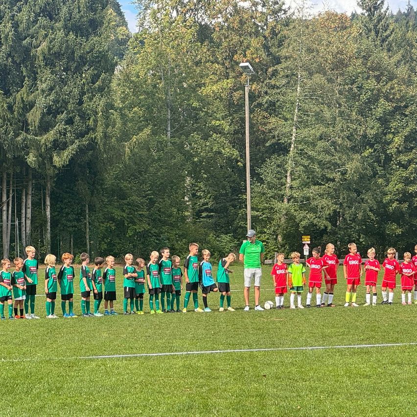 Eine Gruppe junger Fußballspieler in grünen und roten Trikots steht auf einem Feld, ein Trainer in Grün steht neben einem Fußball. Bäume sind im Hintergrund sichtbar.