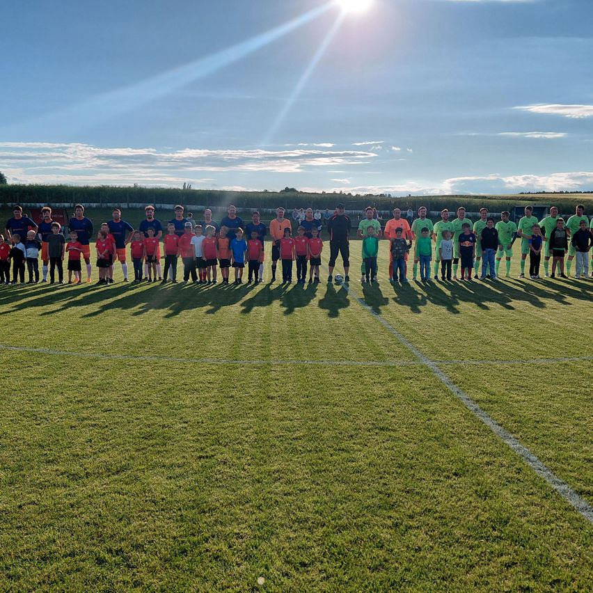 Ein Fußballteam posiert für ein Gruppenfoto an einem sonnigen Tag, geordnet in ihren Uniformen mit einem Rasenfeld im Hintergrund.