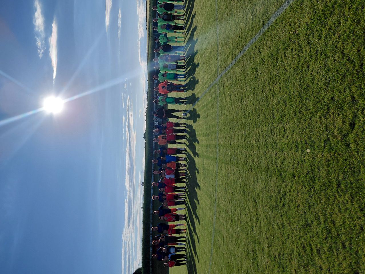Eine Luftaufnahme einer großen Gruppe von Menschen, die sich auf einem Fußballfeld aufstellen. Ihre Schatten werden auf den Rasen geworfen. Die Sonne ist am Himmel sichtbar.