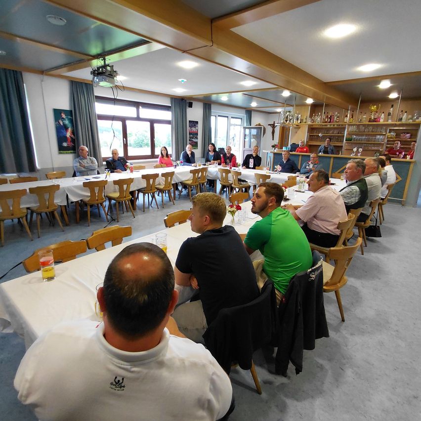 Eine Gruppe von Menschen sitzt um Tische in einem Esszimmer. Ein Mann trägt ein weißes Shirt mit einem Logo. Sie alle schauen nach vorne. Auf den Regalen hinter ihnen befinden sich Flaschen und Trophäen.