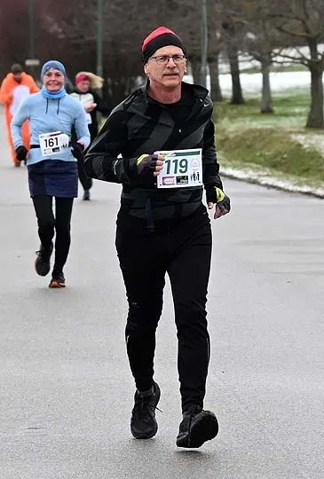Ein Mann in einer schwarzen Jacke läuft bei einem Marathon, mit der Nummer 119 auf seinem Shirt. Hinter ihm läuft eine Frau mit der Nummer 161. Die Straße hat Bäume und Schnee an den Seiten.