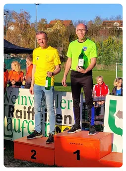 Zwei Männer stehen auf einem Podium und halten Flaschen Wein, einer in Gelb, der andere in Grün, und lächeln für ein Foto. Hinter ihnen steht ein Schild mit 'Weinverkostung'. Im Hintergrund sind Menschen, ein Fußballfeld und ein Zaun.