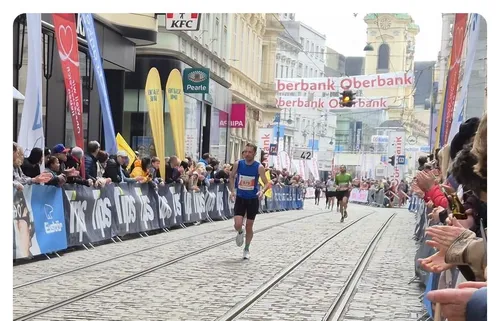 Ein Marathonläufer führt das Rennen auf einer Stadtstraße an, während Zuschauer an der Seite jubeln. Gebäude mit Bannern säumen die Straße.