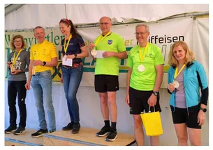 Fünf Personen stehen auf einem Podium. Sie tragen Medaillen, Turnschuhe und halten Papiere. Sie lächeln und schauen in die Kamera. Eine gelbe Papiertüte wird von einer Person gehalten.